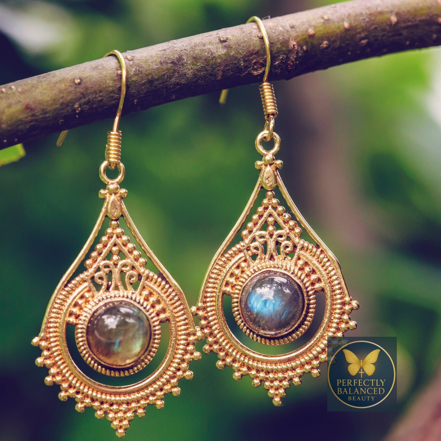 Gold teardrop earrings with intricate detailing and gemstone center displayed on a natural branch — faith-inspired jewelry symbolizing elegance, strength, and divine beauty.